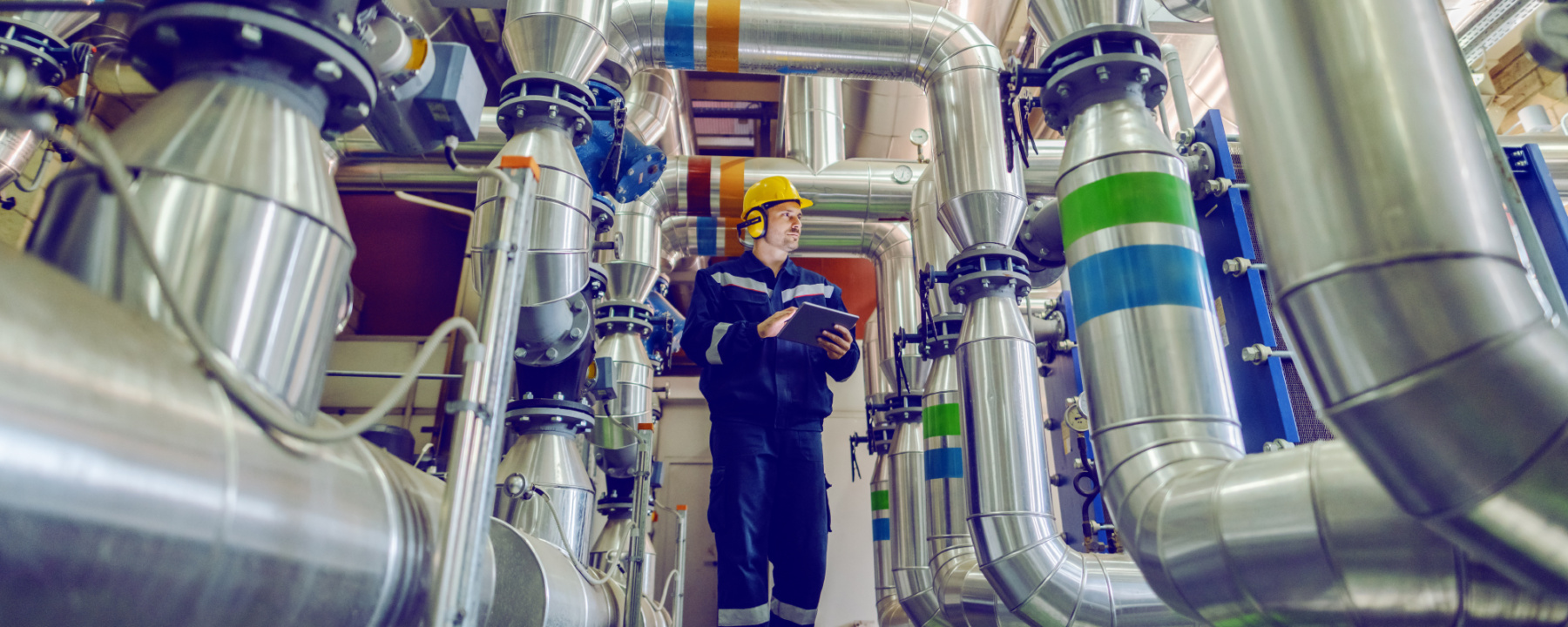 Full length of worker dressed in working clothes, with helmet and antiphons walking around factory and holding tablet
